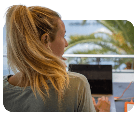 Chica de espaldas tecleando en un portátil, en un balcón mirando a la playa